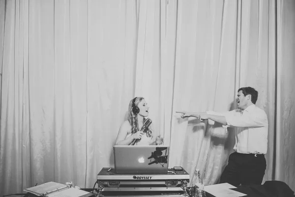 A DJ performing at an event with a laptop and microphone, interacting with a guest.