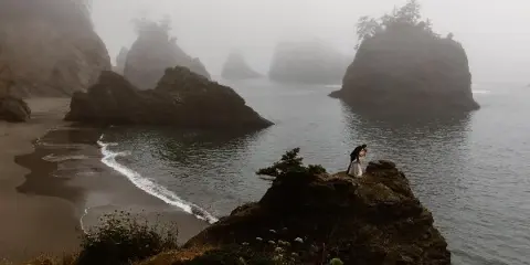 A wedding couple embracing on a rocky outcrop overlooking a misty, dramatic coastline.