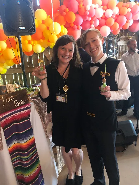 Two staff members wearing Sunrise Specialty Catering vests holding drinks at a decorated event venue.
