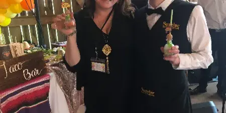 Two staff members wearing Sunrise Specialty Catering vests holding drinks at a decorated event venue.