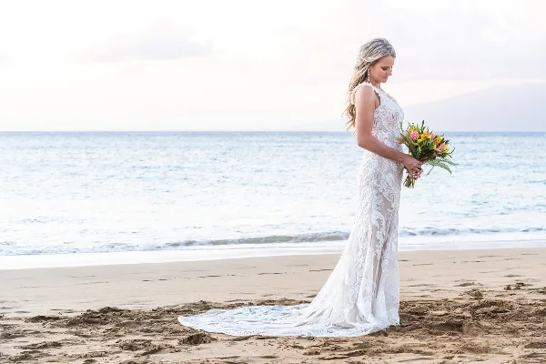 A bride in a white lace gown standing on a sandy beach holding a bouquet of flowers.