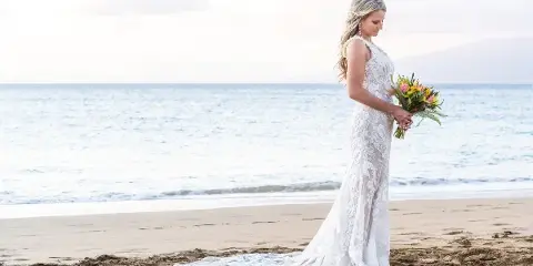 A bride in a white lace gown standing on a sandy beach holding a bouquet of flowers.