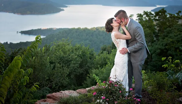 A bride and groom kissing on a scenic overlook with a lake and forest in the background.