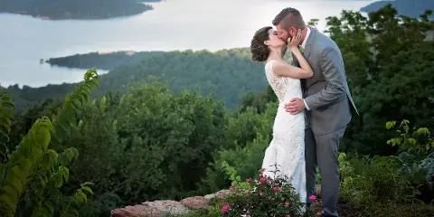 A bride and groom kissing on a scenic overlook with a lake and forest in the background.