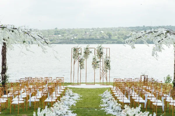 Outdoor wedding ceremony setup with a floral arch, gold chairs, and white floral aisle decorations.