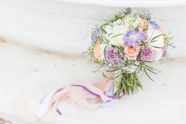 A bridal bouquet with peach and purple roses, white hydrangeas, and greenery resting on a white stone surface.