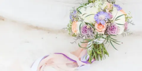 A bridal bouquet with peach and purple roses, white hydrangeas, and greenery resting on a white stone surface.