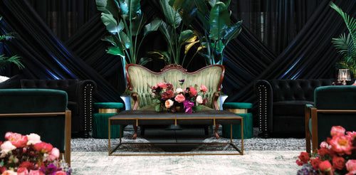 A styled lounge area featuring a vintage upholstered chair, green velvet seating, a wooden coffee table, and floral arrangements.