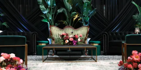 A styled lounge area featuring a vintage upholstered chair, green velvet seating, a wooden coffee table, and floral arrangements.