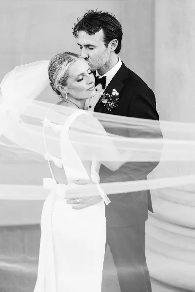 A black and white portrait of a bride and groom sharing a tender moment, with the bride's veil flowing in the foreground.