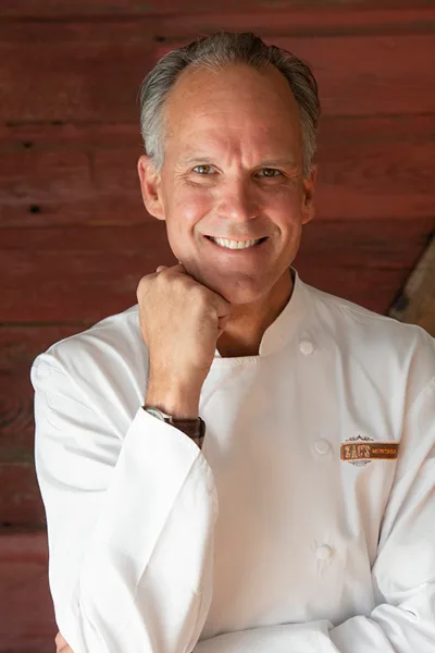 A smiling man in a white chef's coat with a 'Zac's Montana Kitchen' patch, posing against a wooden background.
