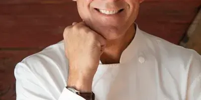 A smiling man in a white chef's coat with a 'Zac's Montana Kitchen' patch, posing against a wooden background.
