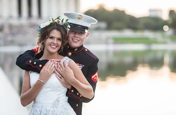 A bride and groom in formal wedding attire, with the groom in a military dress uniform, standing together outdoors.