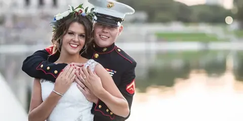 A bride and groom in formal wedding attire, with the groom in a military dress uniform, standing together outdoors.