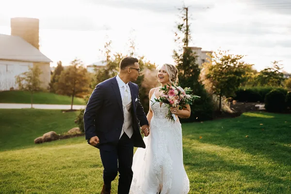 A bride and groom walking hand-in-hand on a grassy lawn at a farm venue during golden hour.
