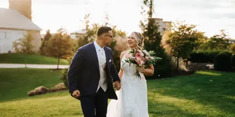 A bride and groom walking hand-in-hand on a grassy lawn at a farm venue during golden hour.