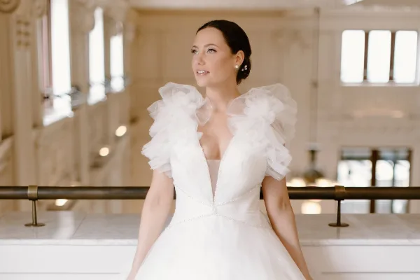 A bride in a white gown with ruffled sleeves, featuring a sleek updo hairstyle and natural makeup, standing in an elegant interior.