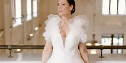 A bride in a white gown with ruffled sleeves, featuring a sleek updo hairstyle and natural makeup, standing in an elegant interior.