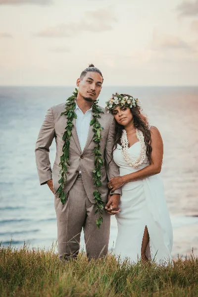A wedding couple standing on a grassy cliff overlooking the ocean, wearing leis and formal wedding attire.