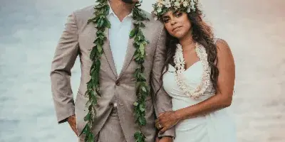 A wedding couple standing on a grassy cliff overlooking the ocean, wearing leis and formal wedding attire.