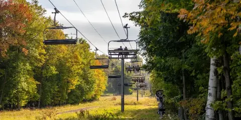 A chairlift carrying passengers up a wooded slope with autumn foliage.