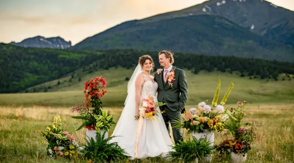 A bride and groom standing in a grassy field with mountains in the background, surrounded by large floral arrangements and holding a bouquet.