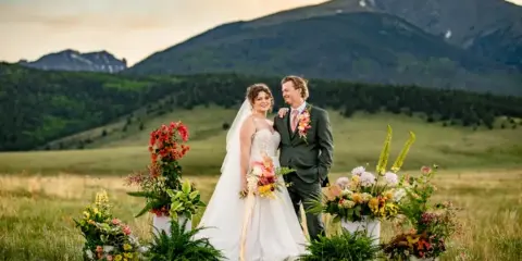A bride and groom standing in a grassy field with mountains in the background, surrounded by large floral arrangements and holding a bouquet.