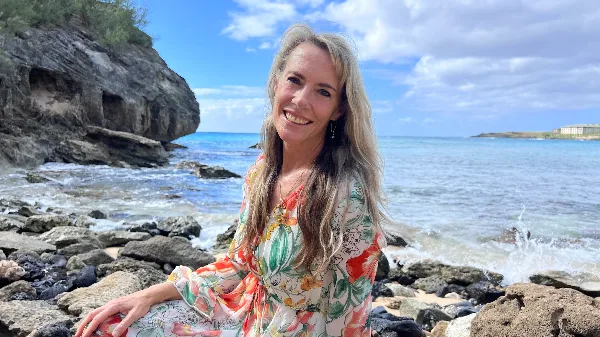 Portrait of a smiling woman with long blonde hair sitting on a rocky beach with the ocean in the background.