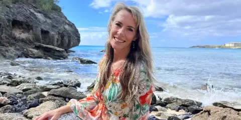 Portrait of a smiling woman with long blonde hair sitting on a rocky beach with the ocean in the background.