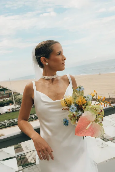 A bride in a white dress and veil with a sleek updo hairstyle and natural makeup, holding a bouquet on a balcony overlooking a beach.