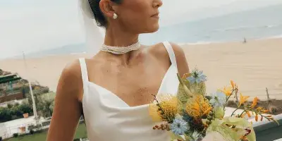 A bride in a white dress and veil with a sleek updo hairstyle and natural makeup, holding a bouquet on a balcony overlooking a beach.