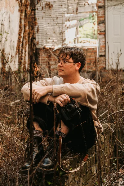 A young man with glasses sits outdoors holding a camera, photographed in a rustic setting.