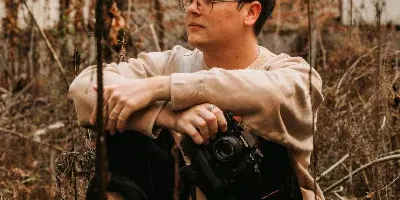 A young man with glasses sits outdoors holding a camera, photographed in a rustic setting.