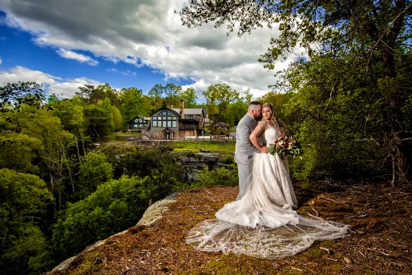 A bride and groom standing on a rocky outcrop overlooking the Burns Bluff lodge and surrounding forest.