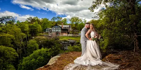 A bride and groom standing on a rocky outcrop overlooking the Burns Bluff lodge and surrounding forest.