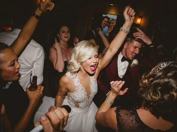 A bride and groom dancing enthusiastically at a wedding reception surrounded by guests.