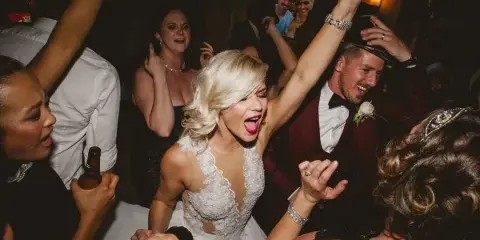 A bride and groom dancing enthusiastically at a wedding reception surrounded by guests.