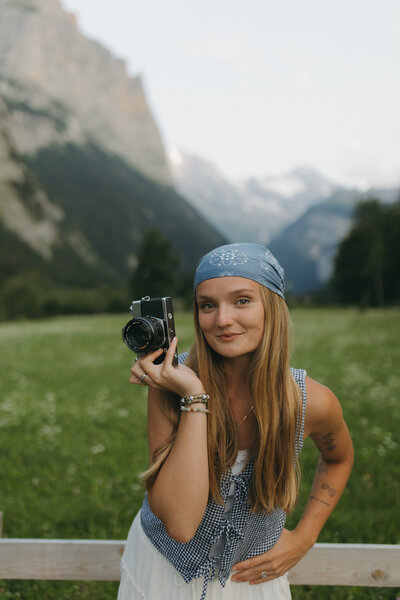 A woman holding a camera in a grassy field with mountains in the background.