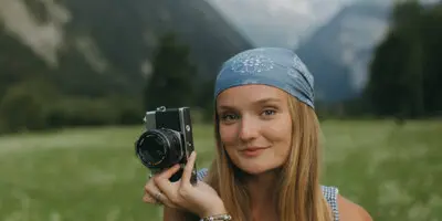 A woman holding a camera in a grassy field with mountains in the background.