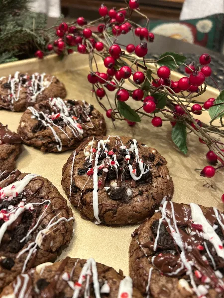 A close-up photograph of chocolate chip cookies topped with chocolate and white icing drizzles, arranged on a tray with red berry decorations.