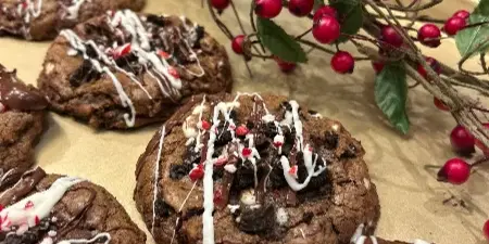 A close-up photograph of chocolate chip cookies topped with chocolate and white icing drizzles, arranged on a tray with red berry decorations.