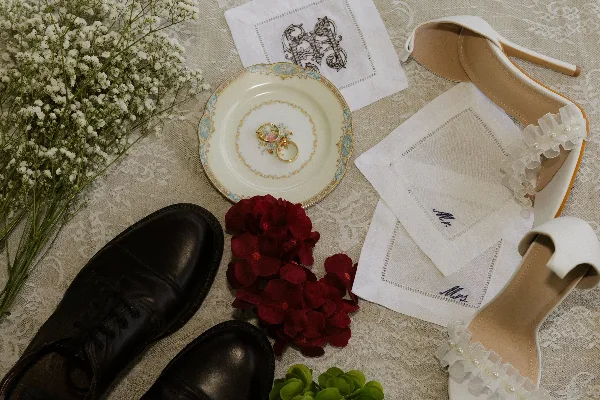 A flat-lay photograph of wedding details including a white flower bouquet, wedding rings on a plate, monogrammed handkerchiefs, and formal shoes.