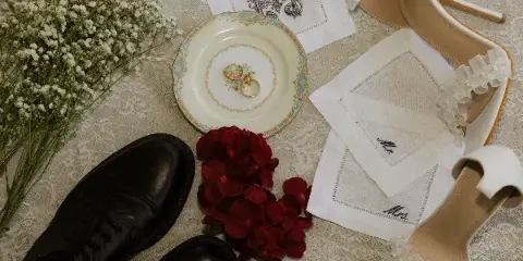 A flat-lay photograph of wedding details including a white flower bouquet, wedding rings on a plate, monogrammed handkerchiefs, and formal shoes.