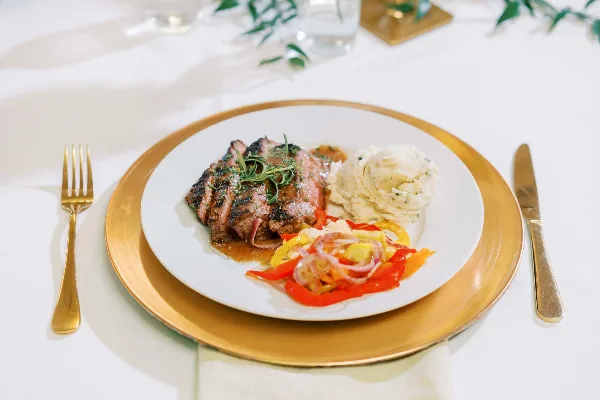A plated meal featuring sliced steak, mashed potatoes, and sautéed vegetables on a white plate with gold charger and cutlery.