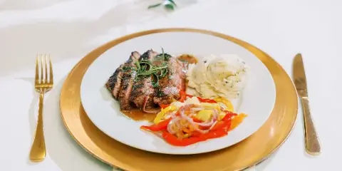 A plated meal featuring sliced steak, mashed potatoes, and sautéed vegetables on a white plate with gold charger and cutlery.