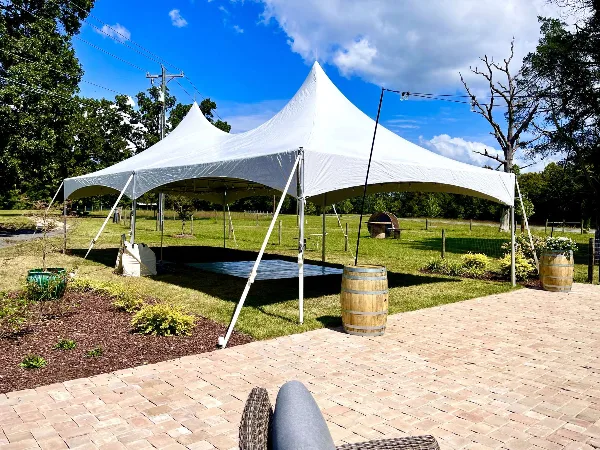 A large white high-peak tent set up on a grassy lawn with a dance floor visible underneath, surrounded by trees and a brick patio.