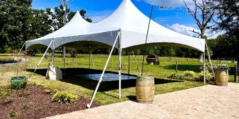 A large white high-peak tent set up on a grassy lawn with a dance floor visible underneath, surrounded by trees and a brick patio.