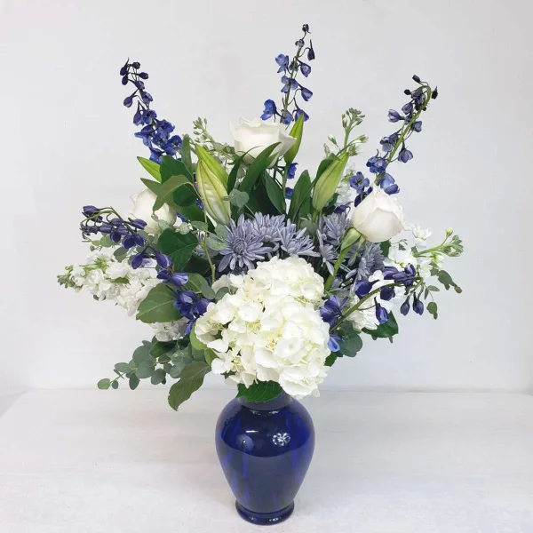 A floral arrangement featuring white roses, hydrangeas, and blue delphiniums in a cobalt blue vase.
