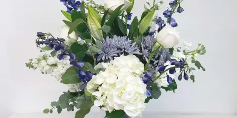 A floral arrangement featuring white roses, hydrangeas, and blue delphiniums in a cobalt blue vase.