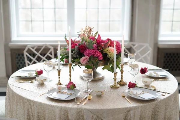 A round wedding table set with a large pink and red floral centerpiece, tall white candles in gold holders, and gold-rimmed glassware.